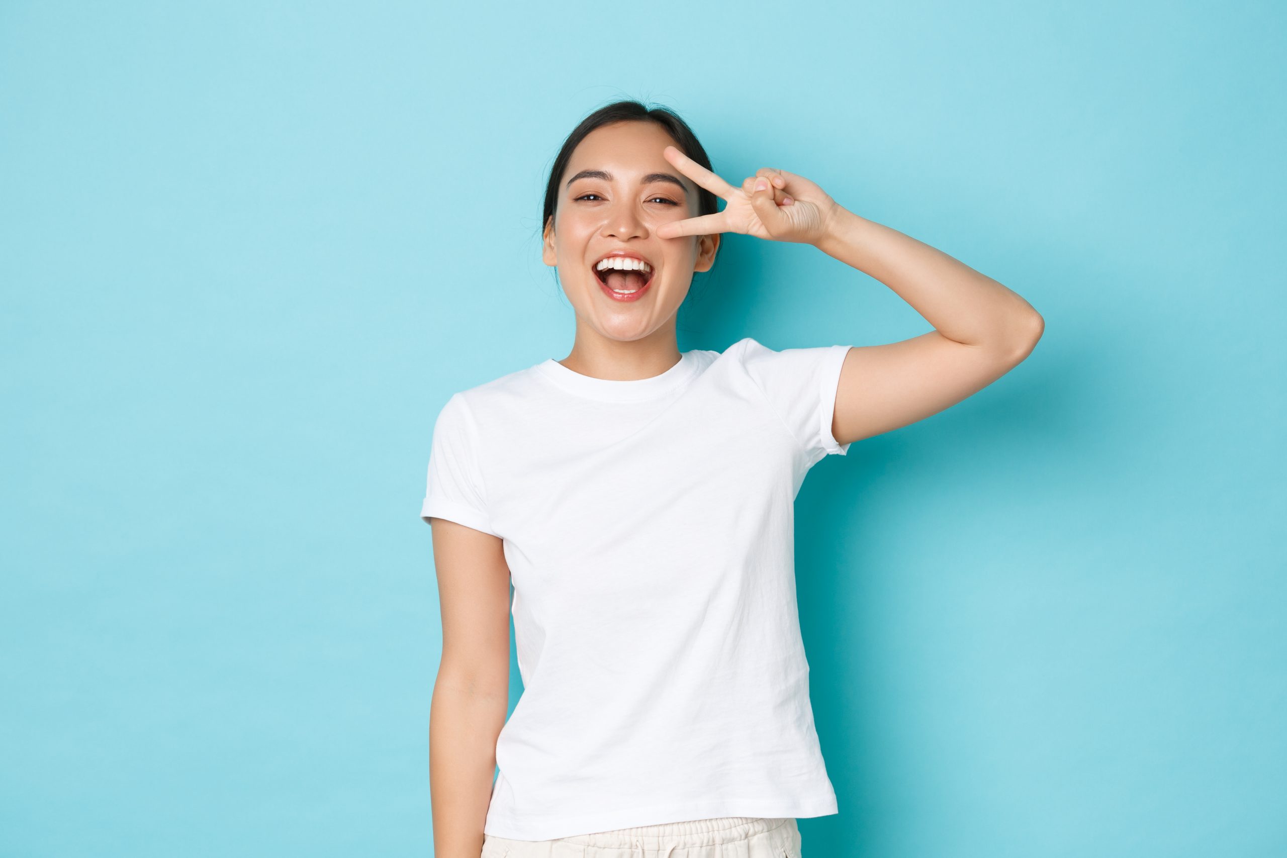 lifestyle, beauty and women concept. cheerful beautiful asian girl in white t shirt looking upbeat and kawaii, showing peace gesture over eye and smiling broadly, standing blue background